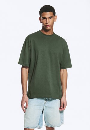 Jeune homme debout, portant un t-shirt oversize vert foncé et un short en jean bleu clair sur un fond blanc uni.