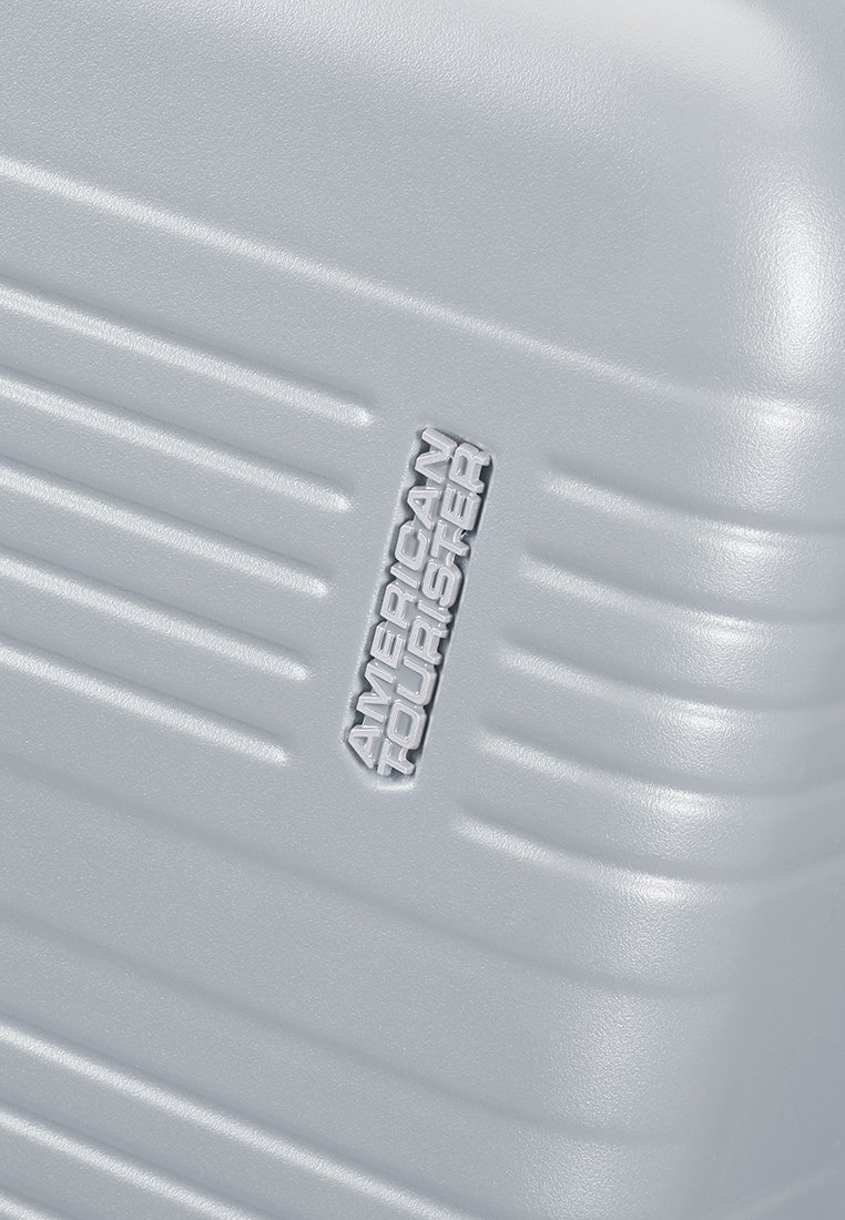 Maleta rígida plateada con superficie texturizada y ranuras verticales. Presenta el logo en relieve "American Tourister" en el costado.