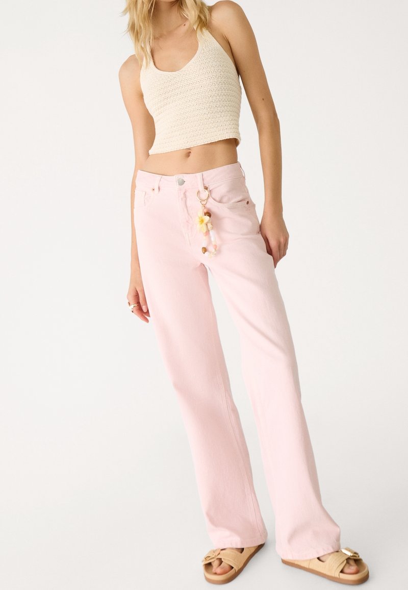 Femme portant un crop top en crochet crème, un pantalon large taille haute rose clair avec un porte-clés, et des sandales ouvertes beiges.