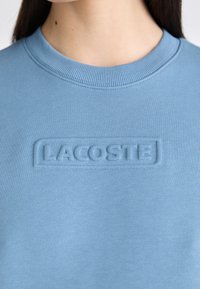Sudadera azul claro con cuello acanalado, parche de logo texturizado que dice "LACOSTE" y un acabado de tela suave.