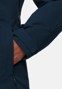 Navy blue jacket cuff with a velcro strap; close-up showing textured fabric, durable material, and a zippered opening at the side.