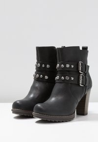 Bottes noires à cheville en cuir synthétique. Elles comportent des clous métalliques et des boucles sur le devant, avec un talon de style bois. Semelle plate.