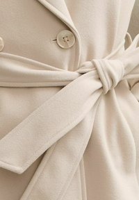 Abrigo cruzado de color crema hecho de tela suave, con botones cruzados, cinturón atado y textura lisa.