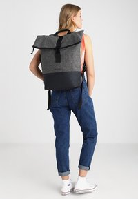 Zaino in tessuto grigio con chiusura a roll-top, sezione inferiore nera e spallacci regolabili. Presenta un design minimalista con una fibbia resistente.