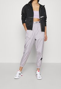 Svart zip-up hoodie med silverdetaljer, kombinerat med ljusgrå joggingbyxor med elastisk midja och cargofickor samt en svart logo. Vita sneakers.