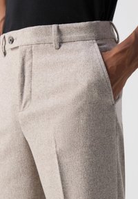 Beige tailored pants with side pockets, textured fabric, and a button closure; features belt loops and a straight leg cut.