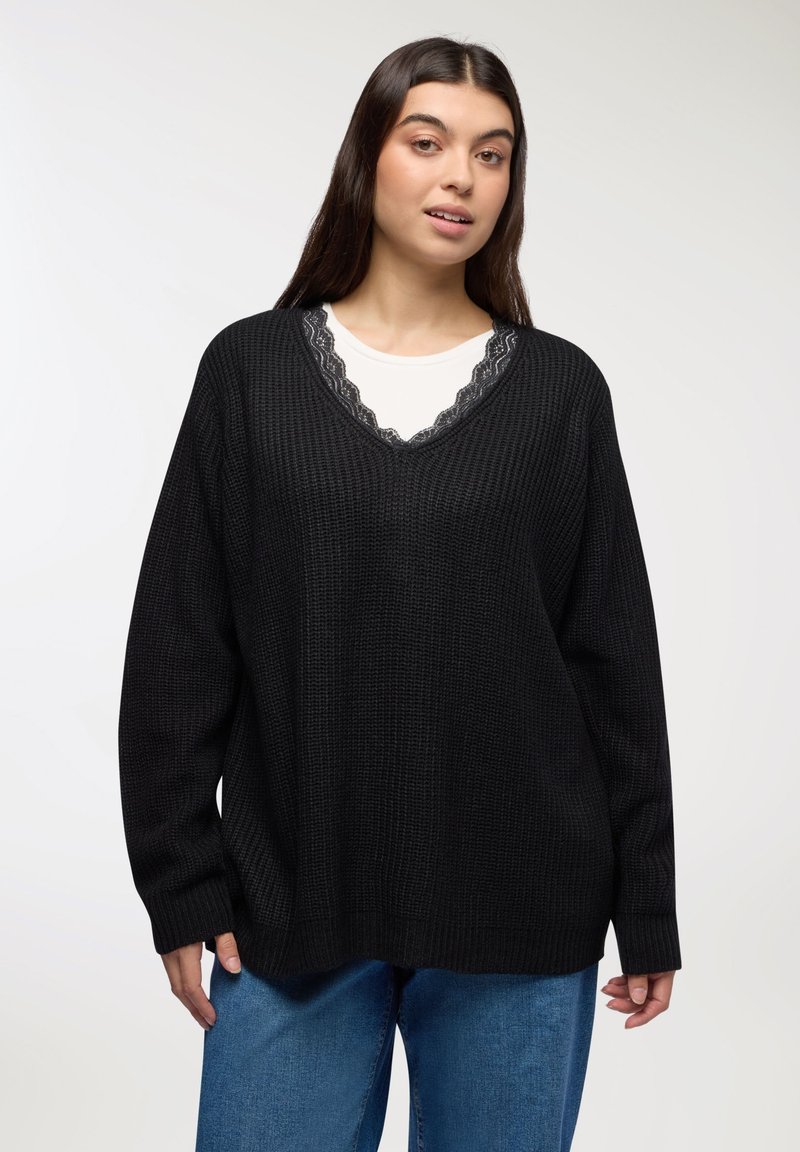 Młoda kobieta z długimi, ciemnymi włosami, ubrana w czarny sweter w serek na białej koszuli z koronkowym wykończeniem oraz niebieskich dżinsach, stojąca na jednolitym tle.