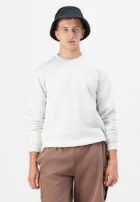Felpa grigia con maniche lunghe e scollatura rotonda, abbinata a pantaloni marroni. Il modello indossa un cappello a secchiello nero.