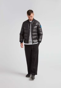 Giacca puffer nera con motivo trapuntato, chiusura con zip e polsini a coste; abbinata a un top grigio e pantaloni neri; design minimalista, senza loghi visibili.