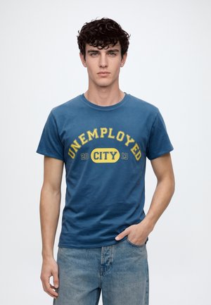 Un jeune homme présente un T-shirt bleu avec l’inscription jaune « UNEMPLOYED CITY » et un jean sur un fond blanc uni.