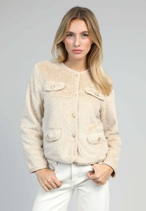 Veste texturée beige avec un col rond, fermeture à boutons sur le devant et deux poches poitrine avec des boutons dorés. Portée avec un pantalon blanc.
