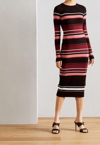 Robe en maille côtelée à rayures noires, bordeaux et roses, avec manches longues et ourlet arrivant au genou. Présente des accents horizontaux blancs.