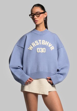Light blue knitted sweater with dropped shoulders, featuring white text "WRSTBHVR 030." Worn over a light textured skirt, with ribbed cuffs.