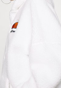 White fleece jacket with a quarter zip, featuring an embroidered logo in orange, navy, and white. Soft texture and relaxed fit.