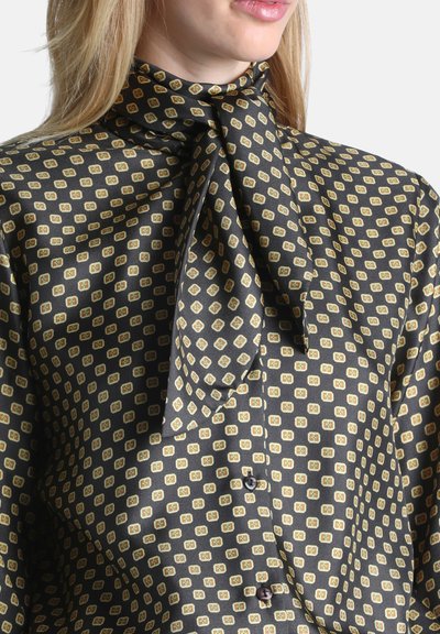 Femme portant une blouse noire avec un motif carré jaune et vert et un col haut noué en nœud papillon, montrant le bas du visage et les cheveux blonds.