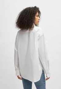 White, long-sleeve shirt with a relaxed fit, side slits, and ties at the cuffs. Smooth texture and slightly curved hem. Worn with blue jeans.