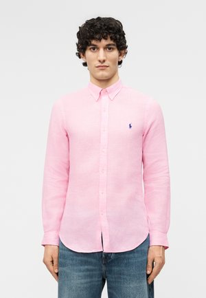 Jeune homme aux cheveux bouclés portant une chemise rose clair à boutons avec un petit logo bleu et un jean bleu, sur un fond uni.