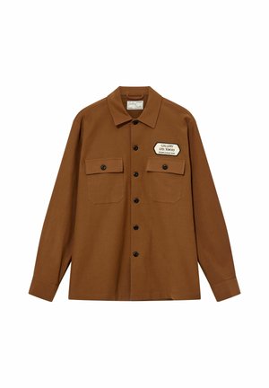 Brun skjorte med knapper og to brystlommer samt et hvidt mærke med teksten "Gallery Ltd. Tokyo Studio Collection" på højre lomme.