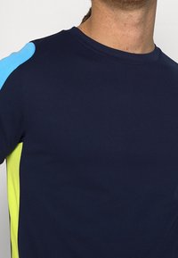 Námořnicky modré sportovní tričko se světle modrými a jasně žlutými akcenty na ramenou, vyrobené z hladké, texturované látky. Kulatý výstřih.