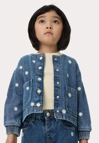 Giubbotto di denim con ricami di margherite bianche, bottoni a pressione metallici e una vestibilità ampia. Presenta polsini elastici e tasche laterali. Indossato con una maglietta color crema.