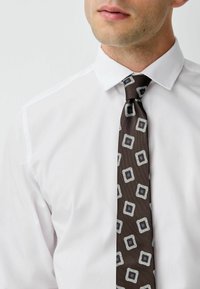 Brown tie with a textured surface, featuring a geometric pattern of squares in light silver and blue tones, worn over a white shirt.