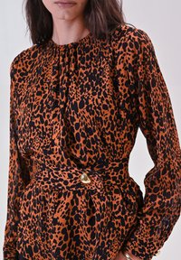 Blusa a stampa leopardata in arancione e nero, scollo arricciato, maniche lunghe, vita aderente con dettaglio di bottone dorato. Tessuto liscio e leggero.