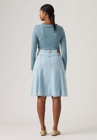 Γυναίκα με πλεξούδες, που φοράει μπλε μπλούζα με μακριά μανίκια, ανοιχτόχρωμη φούστα από τζιν μέχρι το γόνατο και χρυσά παντόφλες, στέκεται με την πλάτη προς τα εμπρός.