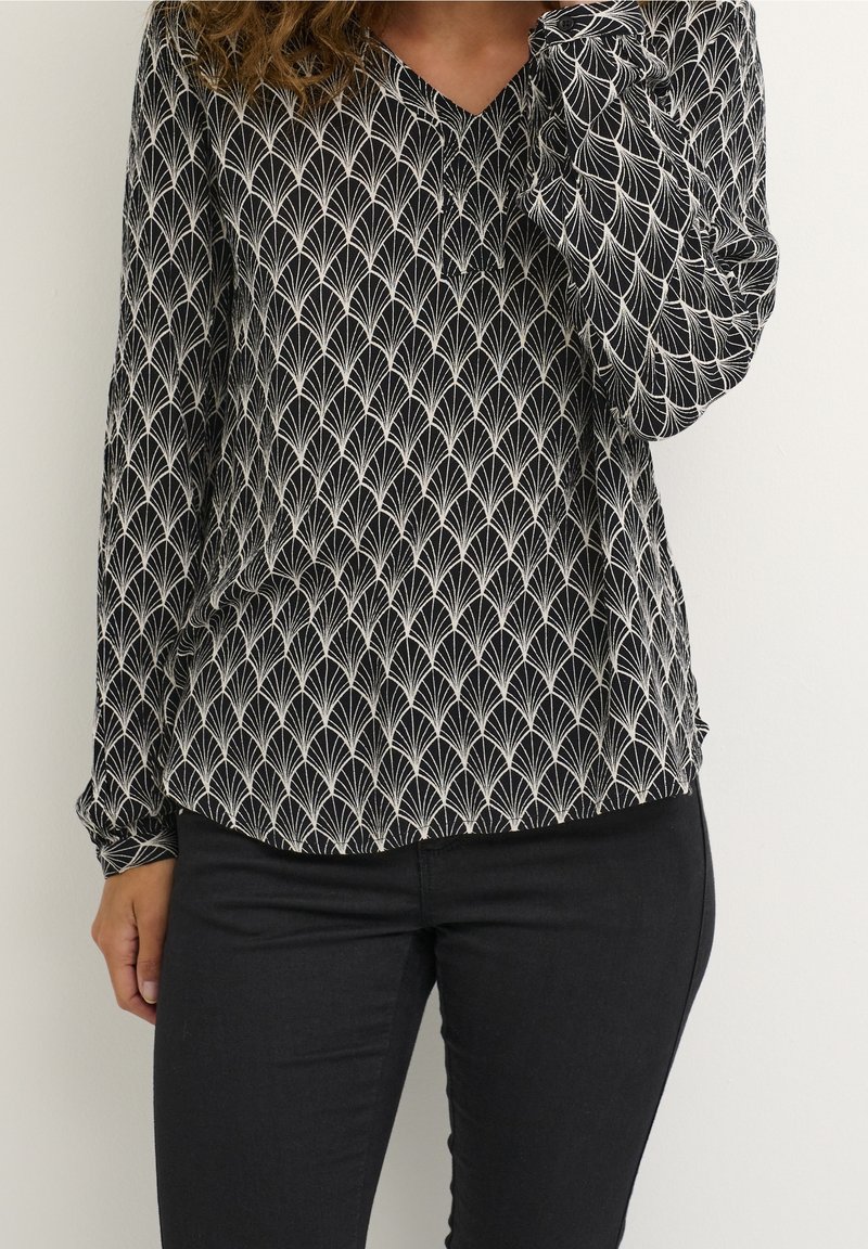 Femme portant un chemisier à manches longues noir et blanc à motif avec col en V et un pantalon noir ajusté, debout devant un fond uni.
