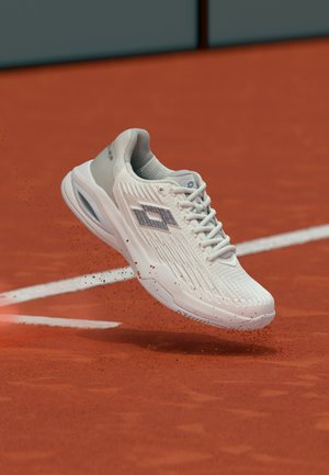 Zapato deportivo blanco con logo gris en el aire sobre una pista de tenis de arcilla roja con líneas blancas, partículas de tierra dispersándose debajo de la suela.