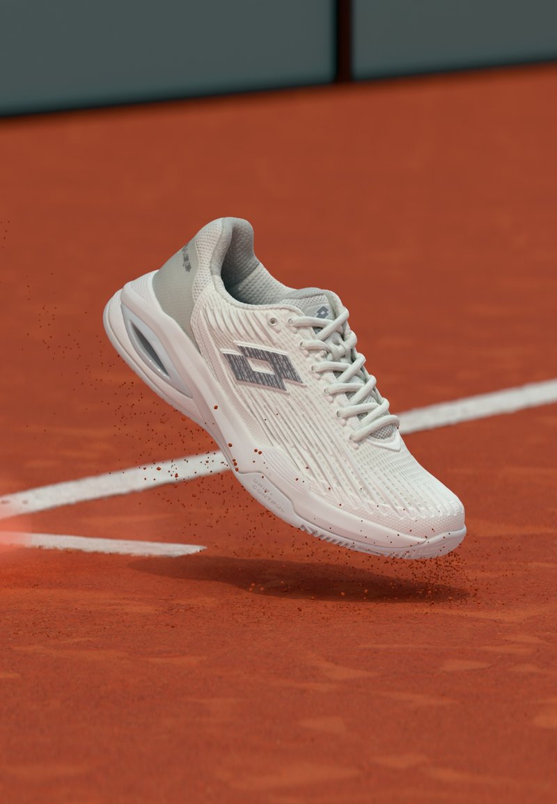 Biela športová obuv so sivým logom vo vzduchu nad červeným antukovým tenisovým kurtom s bielymi čiarami, pod podrážkou sa rozptyľujú čiastočky špiny.