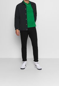 Polo verde sotto una giacca leggera nera, pantaloni slim-fit neri e sneaker bianche con dettagli neri. Design semplice, look casual.