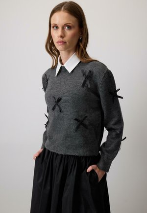 Maglione grigio con dettagli in nastro nero sulla parte anteriore, scollo rotondo e maniche lunghe, abbinato a una gonna nera voluminosa.