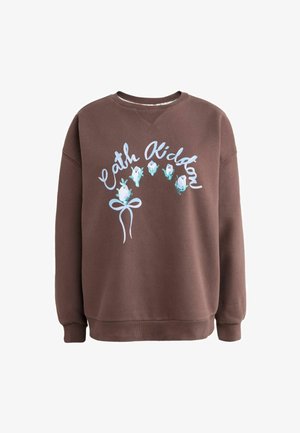 Kobieta w brązowej bluzie Cath Kidston z kwiatowym wzorem, białej koszuli, podwiniętych ciemnych dżinsach i brązowych tenisówkach, stojąca w pomieszczeniu.