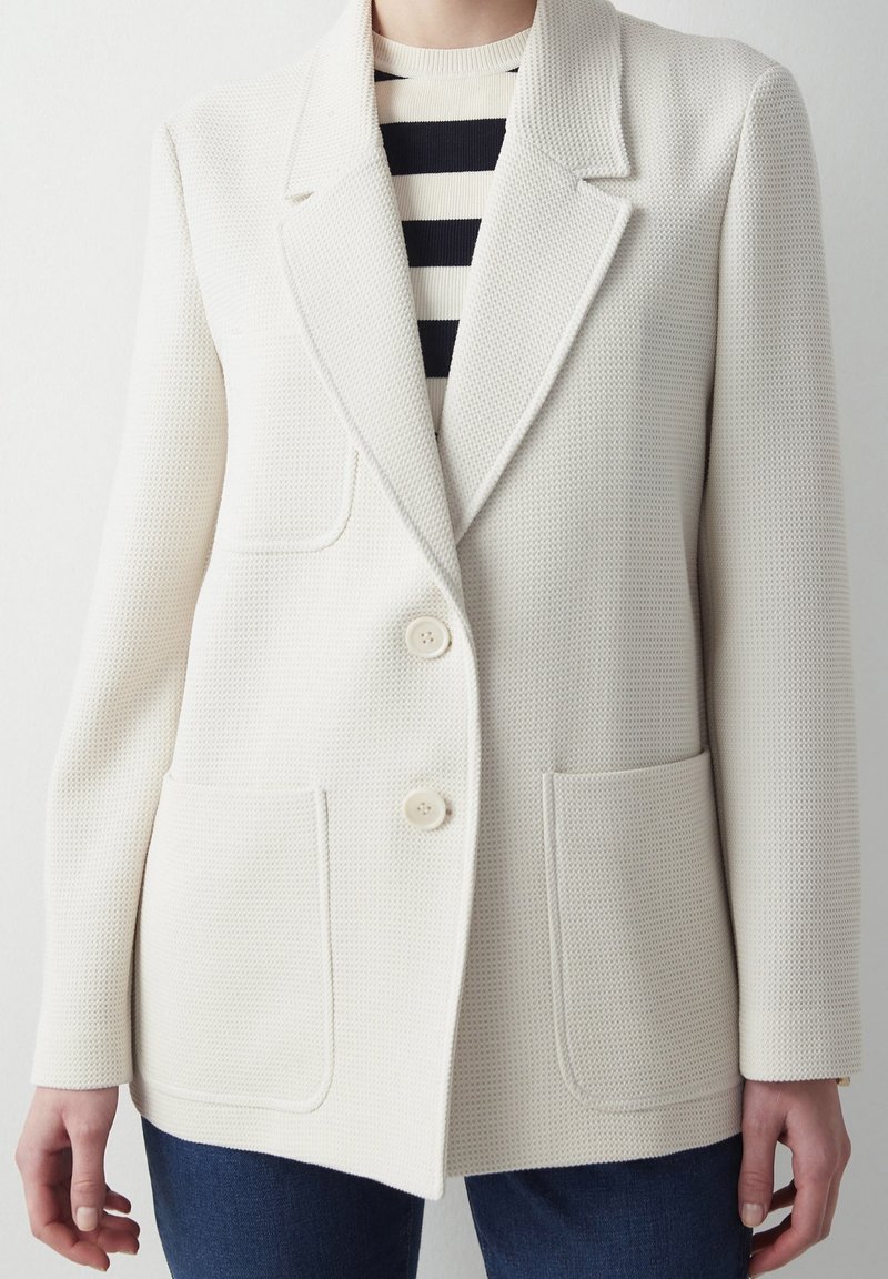 Blazer blanc texturé avec deux poches avant, col à revers cranté et deux boutons. Associé à un haut rayé noir et blanc en dessous.
