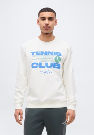 Νέος άνδρας που φοράει λευκό φούτερ με το κείμενο "Tennis Club" και γραφικό με μπάλα του τένις, συνδυασμένο με σκούρο γκρι παντελόνι φόρμας.