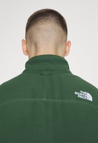 Mężczyzna o krótkich ciemnych włosach widziany od tyłu, ubrany w zieloną polarową kurtkę The North Face z logo na górnej części pleców.