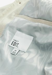 Lichtbeige stoffen interieur met een glanzende zilveren rand en een subtiel patroon. Etiket leest "NEXT EDIT" in zwarte tekst, maat M, gemaakt in Vietnam.