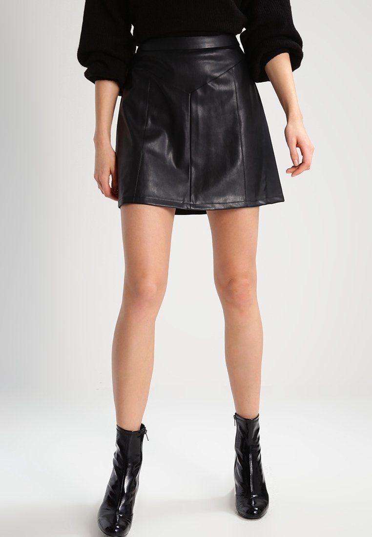 Falda mini de cuero negro con una cintura ajustada, dobladillo ligeramente acampanado y textura suave. Combinada con botas de tobillo negras brillantes.