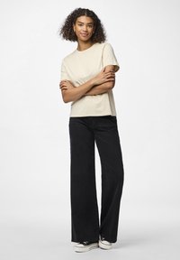 Beige bomulls t-skjorte med korte ermer, kombinert med vide svarte cordbukser. Modellen står med armene krysset iført svarte joggesko.