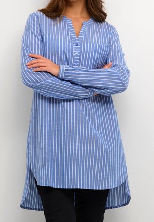 Frau mit verschränkten Armen, die eine blau-weiß vertikal gestreifte, langärmelige Tunika und eine schwarze Hose vor einem schlichten Hintergrund trägt.