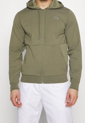 Homme portant un sweat à capuche zippé vert olive avec poches avant et un petit logo crocodile sur la poitrine, associé à un pantalon blanc.