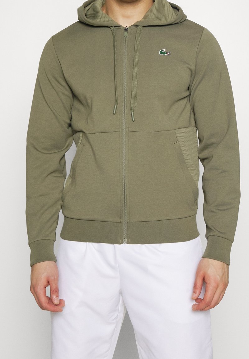 Homme portant un sweat à capuche zippé vert olive avec poches avant et un petit logo crocodile sur la poitrine, associé à un pantalon blanc.