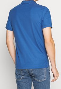 Polo en coton bleu avec col classique, manches courtes et fentes latérales. Texture lisse, design simple, sans motifs ni logos visibles.