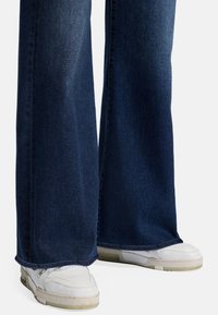 Jeans in denim blu scuro a gamba larga con una texture liscia e orlo pulito, abbinati a scarpe da ginnastica bianche con dettagli sottili.