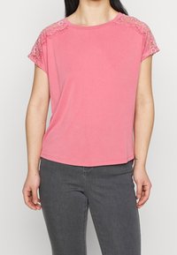 Top rosa a maniche corte realizzato in morbido tessuto con dettagli in pizzo sulle spalle. Collo rotondo e vestibilità comoda, abbinato a pantaloni grigi.