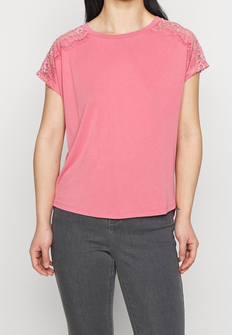 Top rosa a maniche corte realizzato in morbido tessuto con dettagli in pizzo sulle spalle. Collo rotondo e vestibilità comoda, abbinato a pantaloni grigi.