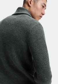 M�ørk grå strikket sweater med et tekstureret, ribbet mønster; har en ombukket krave og lange ærmer. Nærbillede fra ryggen.