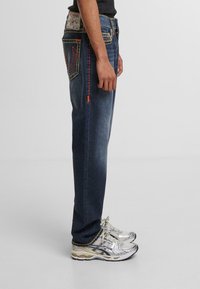 Jeans de denim en índigo oscuro con costuras en contraste. Presentan un corte recto, bolsillos laterales y una etiqueta de la marca en la parte trasera de la cinturilla.