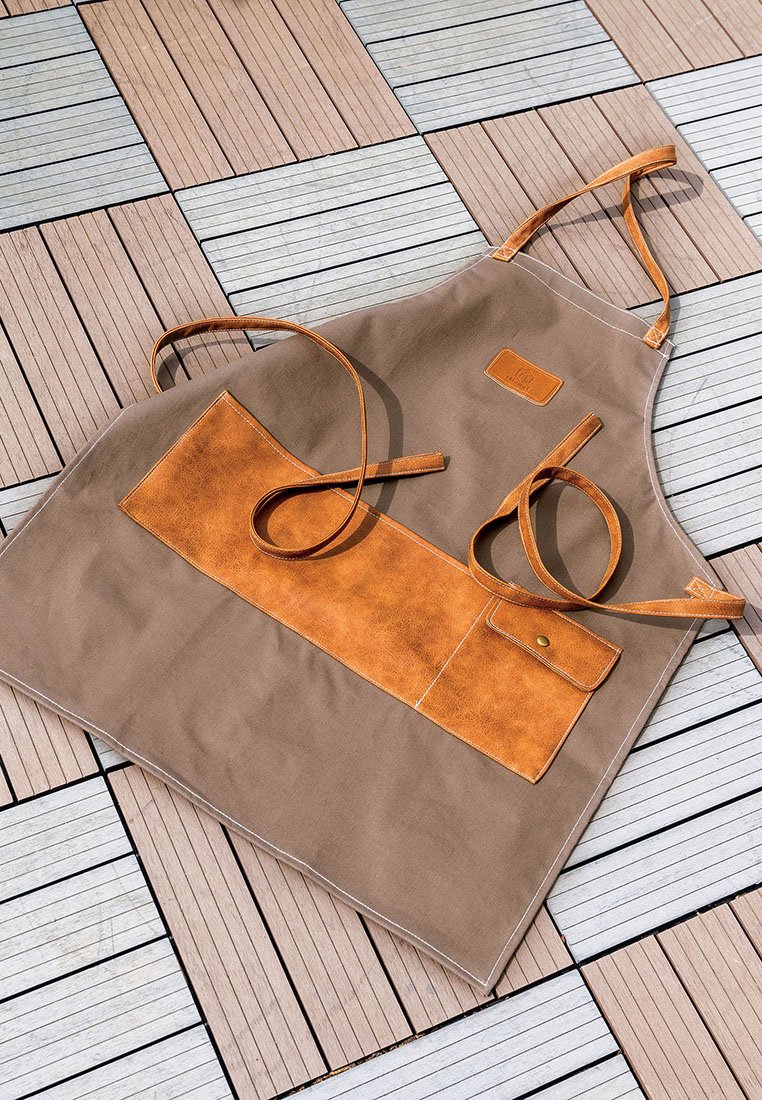 Tablier en toile marron avec une poche en cuir beige, bretelles réglables et un bouton-pression en métal. Texture lisse avec des couleurs contrastées et des détails de couture.