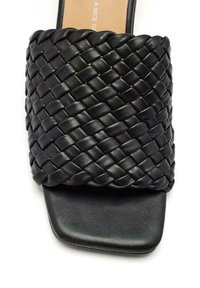 Sandale en cuir tressé noir avec une large sangle, un bout carré et une semelle plate. La texture semble lisse avec un détail en motif tressé.